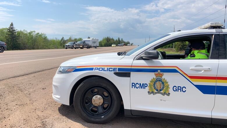 An officer in a fluorescent green vest sits in a white RCMP car at the side of a highway as traffic drives past.