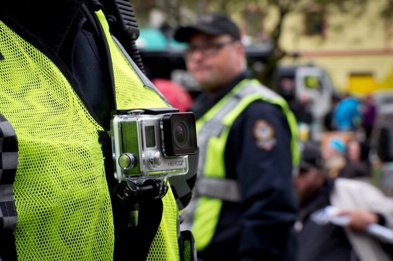 A GoPro camera is seen on the chest of someone wearing a high-visibility vest, with a police officer and a parkgoer in the backgroud.