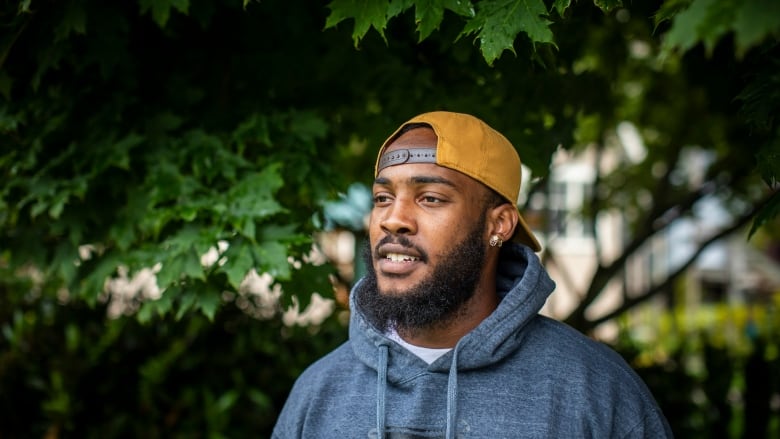 A Black man is pictured in a blue hoodie and yellow baseball cap in front of green trees.