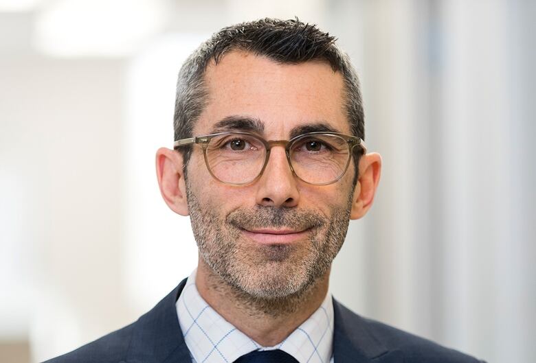 A man with glasses in a suit and tie smiles slightly for a portrait.