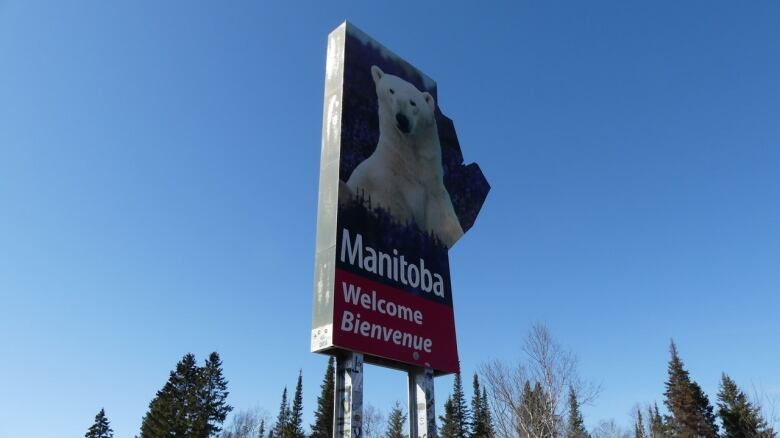 A sign in the shape of the province of Manitoba stands against a clear blue sky. The sign bears the image of a polar bear and says, 