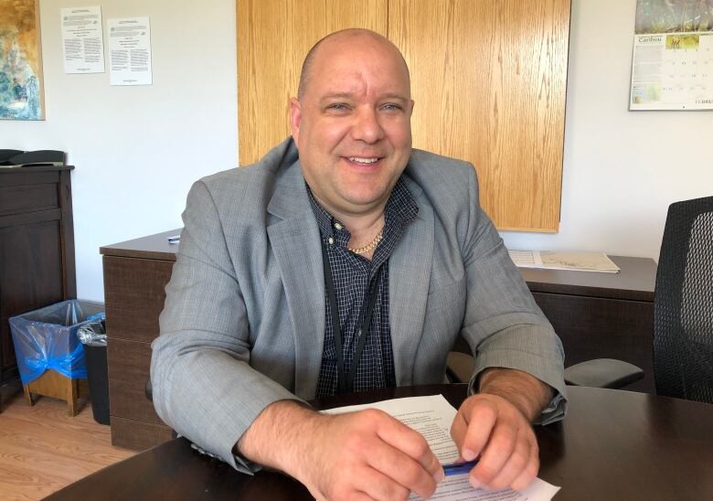 A man in a grey jacket sits at a desk with his hands holding a pen, smiling at the camera.