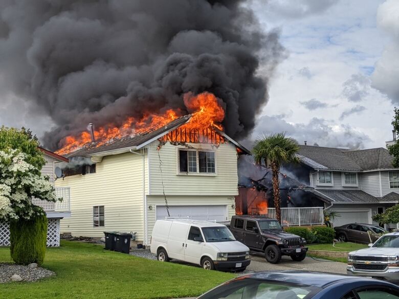 A yellow-sided single-family home with its roof in flames and a white van and a black truck parked in the front driveway. The house is next to a row of other similar homes.