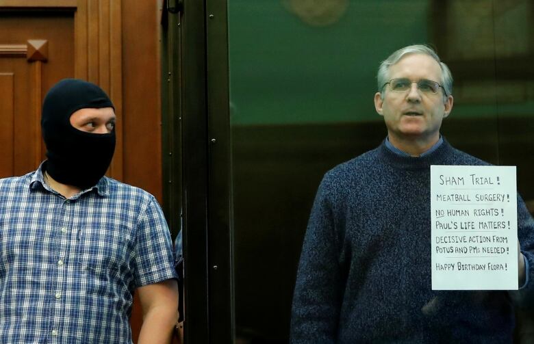 A grey-haired man, wearing a blue sweater and glasses, stands on the right holding up a sign next to a man on the left with a black mask covering his head and face. 