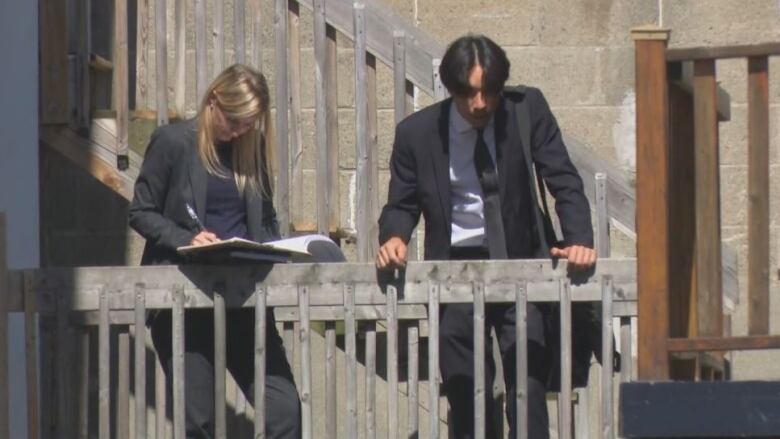 A woman, left, and a man, right, both wearing black suits, leaning on a wooden railing. The woman is writing in a book.