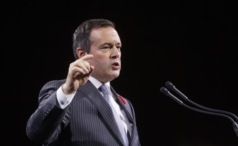 A man in a dark suit speaks in front of microphones.