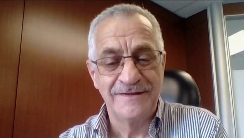 A man with greying hair and a mustache looks down as he faces a camera.