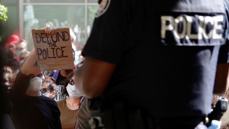 A person in a mask within a crowd holds up a 'defund the police sign' while the back of a police officer is visible