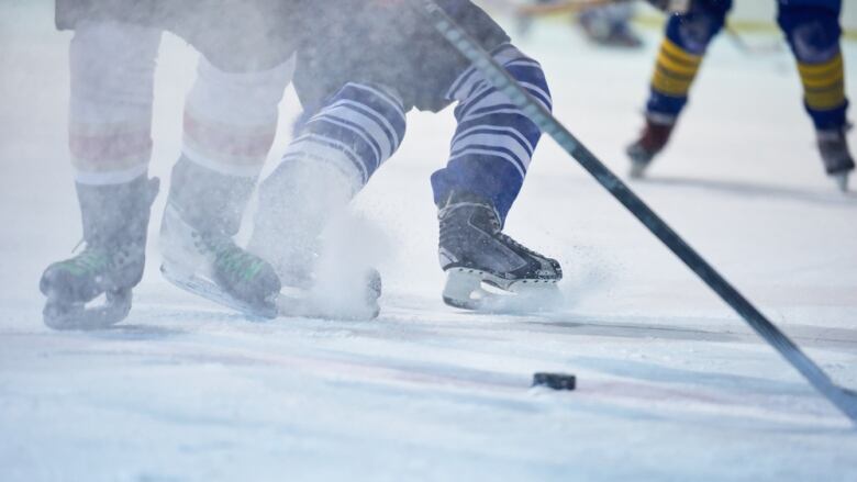 A closeup photo shows the skates of players chasing after a puck on a skating rink.