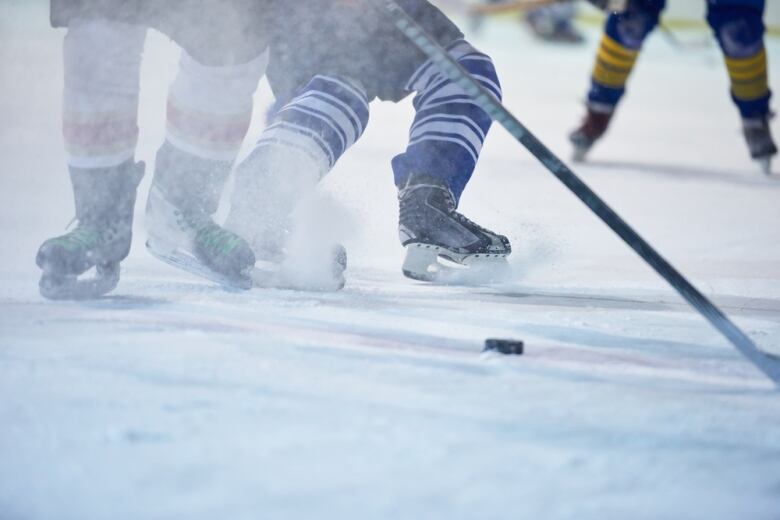 A closeup photo shows the skates of players chasing after a puck on a skating rink.