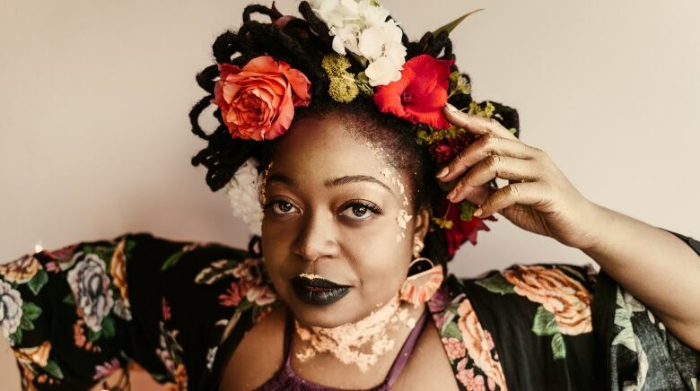 A woman with flowers in her hair has gold flakes on her lips, forehead and neck. She's wearing a floral coat and has one hand holding her head. 