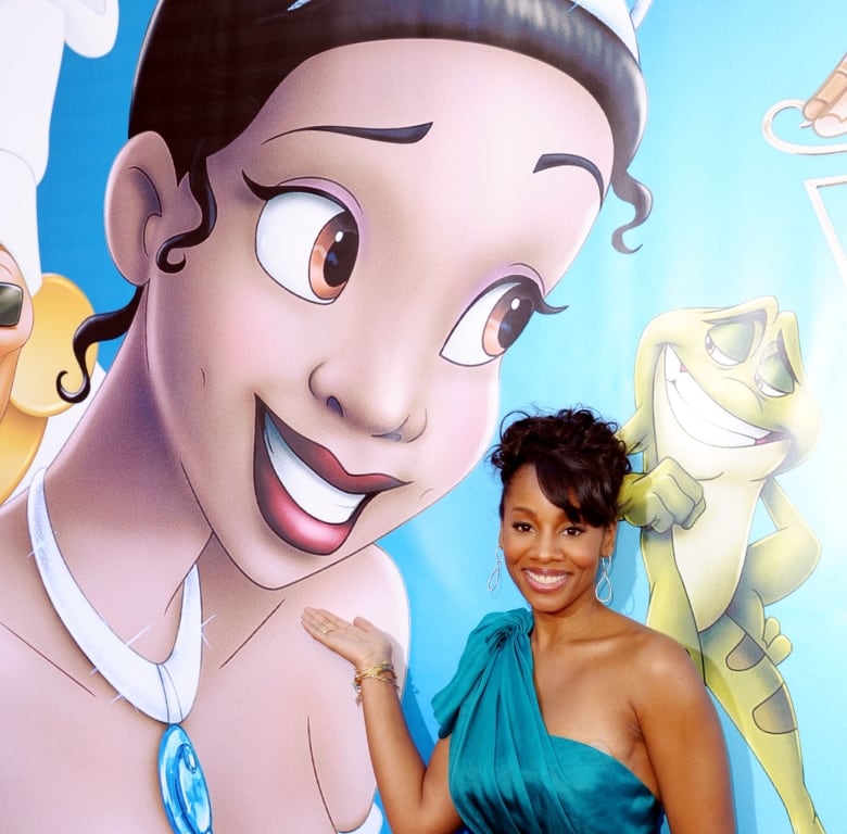 A Black woman smiles as she gestures to a poster of a cartoon character she is playing displayed on a wall behind her. 