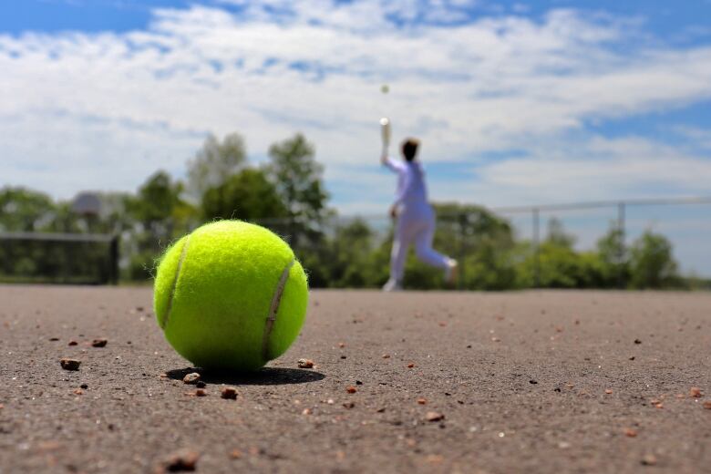 A tennis ball lays on the ground. There's a tennis player in the background.