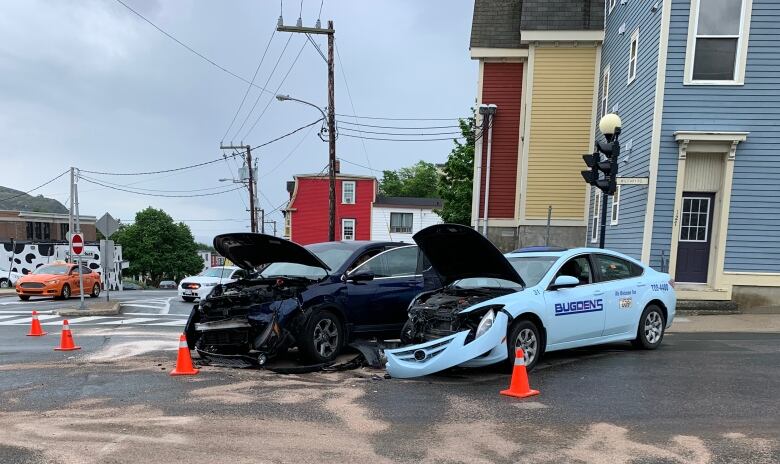 A road with houses behind it and two cars that collided. There are orange pilons around the cars.