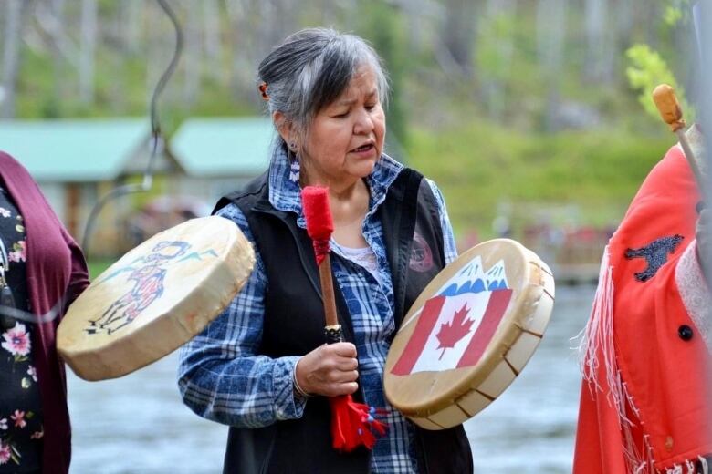 An Indigenous woman plays a drum and sings with standing with other people.