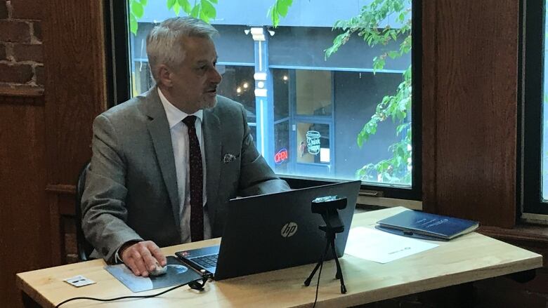 A man with short grey hair sits at a desk in front of a computer and is looking off in the distance to the right of the photo. He is wearing a grey suit, white dress shirt and dark tie.