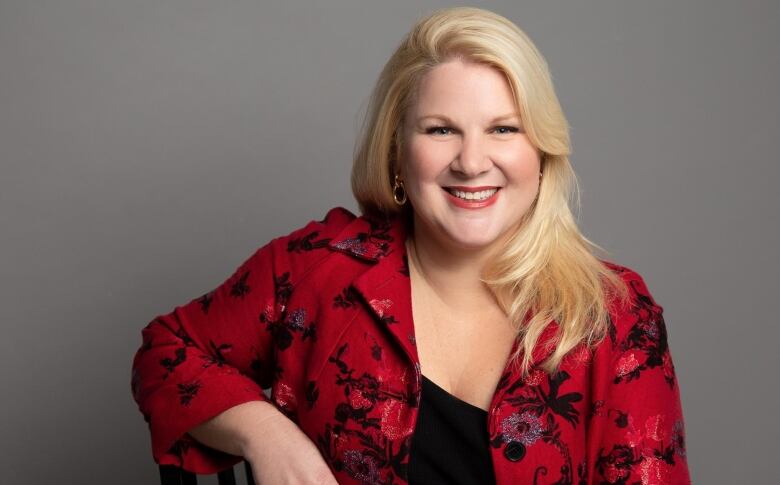 A blonde woman in a red and black jacket looks directly at the camera. 