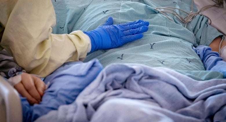 A close up of a person's arm, in a yellow sleeve with a blue latex glove, touching the chest of someone lying on a hospital bed.