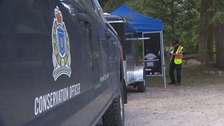 An up-close picture of a B.C. Conservation Officer car, with a man in a high-vis vest in the background.