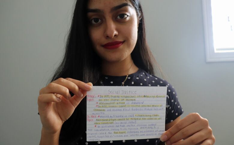 A woman with long dark hair holds a cue-card with writing on it.