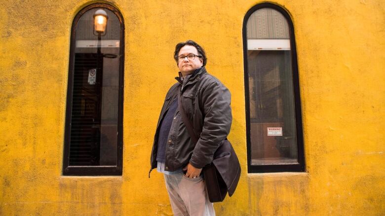 An Anishinaabe man standing away from the camera sideways but looking into the camera, in front of a yellow building with oval windows. He is wearing a bag over his shoulder.