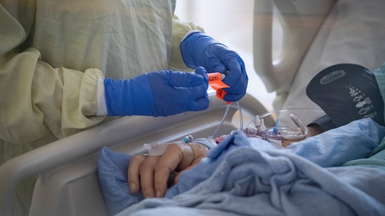 A close-up of a patient's hand as they lay in a medical bed. A health-care worker wearing a gown and gloves attends to them.