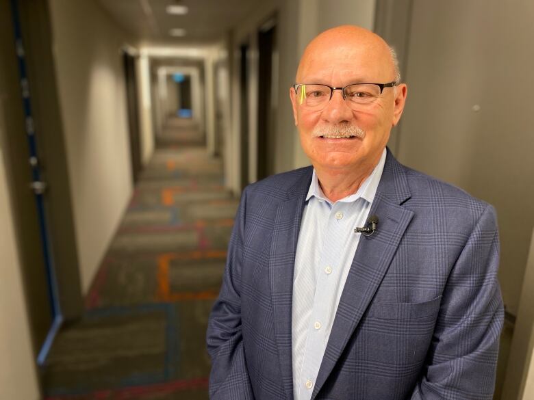 Man wearing glasses, suit jacket and dress shirt stands in a hotel corridor.