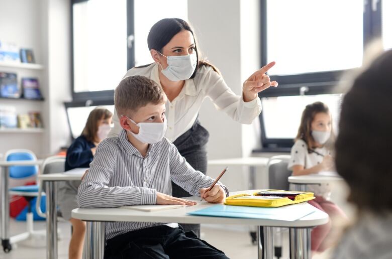 Teacher standing over student pointing to the front of the classroom. Both are wearing facemasks.