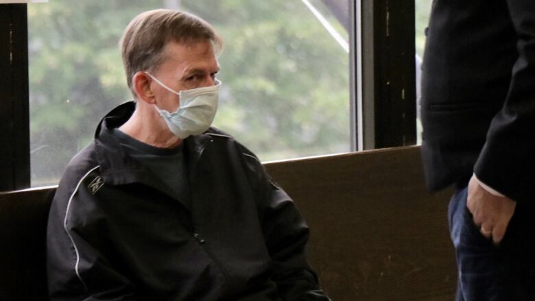 A man wearing a black windbreaker and a surgical face mask sits in a courthouse lobby.