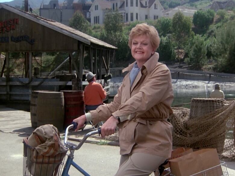 A middle-aged woman with short strawberry blonde hair and a short tan trenchcoat smiles as she pushes an old-fashioned bicycle next to a river.