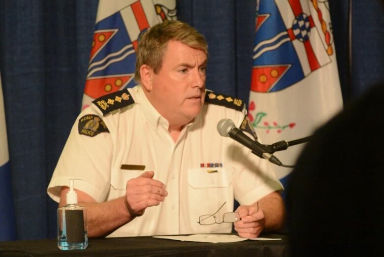 An RCMP officer sits at a table in a dark room, speaking into a microphone.