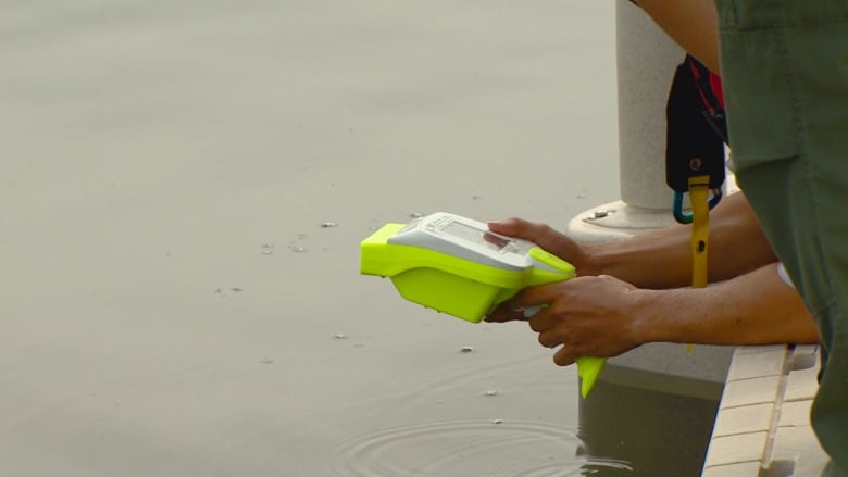 a bright yellow dveice in someones hands, as they lean over the edge of a wharf. 