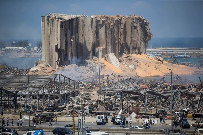 In the background looms a tall building by the water, shredded and covered in debris, surrounded by sand, rubble and large chunks of concrete. In front of it are the charred remnants of destroyed smaller buildings, with nothing left but beams. Dozens of people are milling about near the rubble and on the street in front of it.