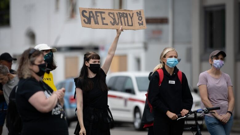 A woman holds up a cardboard sign that reads Safe Supply Now