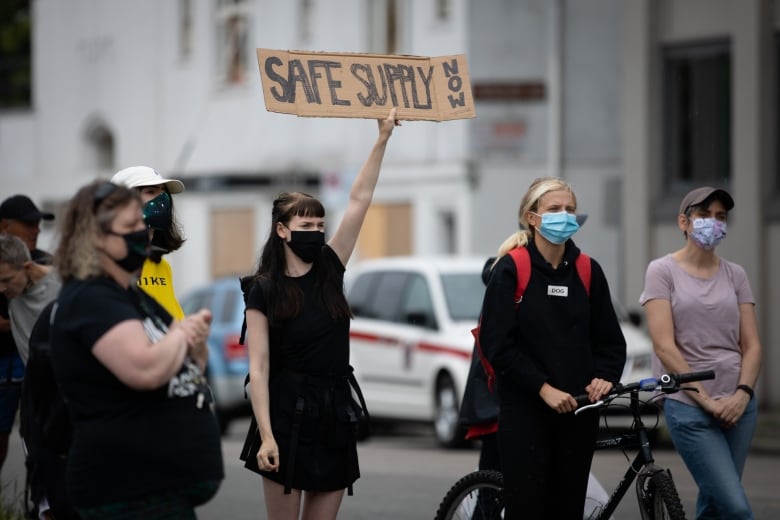 A woman holds up a cardboard sign that reads Safe Supply Now