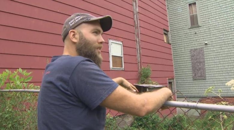 A bearded man wearing a T-shirt and ball cap leans on a fence. 