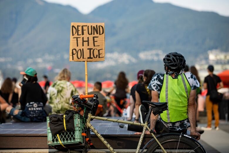 A man sits on a bench, next to a bike with a sign that reads 'Defund the Police'.