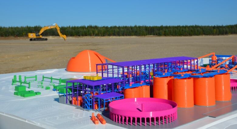 A colourful model of a gold mine sits on a table in a large open area with a piece of heavy equipment and a forest in the background 