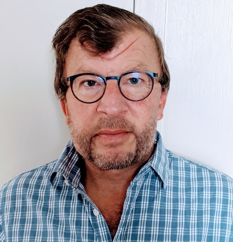 Man in glasses with blue button up stands in front of white wall.