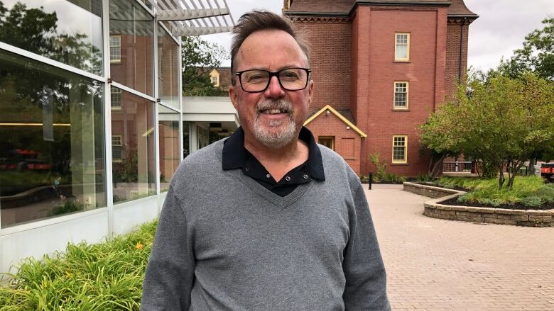 A man with glasses a beard and a blue sweater vest stands on a university campus