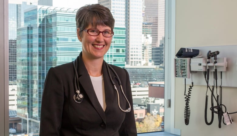 Dr. Christine Luelo stand in front of a window in an examination room. She is wearing a brown suit and has a stethoscope around her neck.