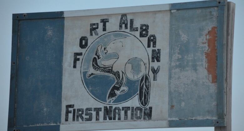 A faded blue and white wooden sign reads 'Fort Albany First Nation' with a picture of a polar bear and a fish