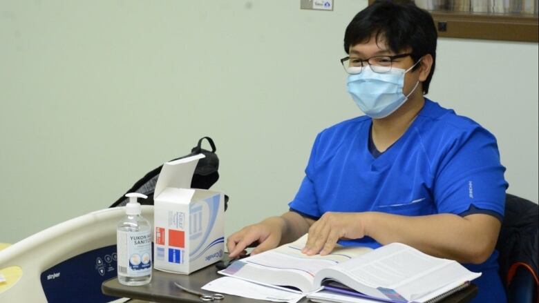 A young man in scrubs and a surgical mask sits in a hospital, flipping through a text book.