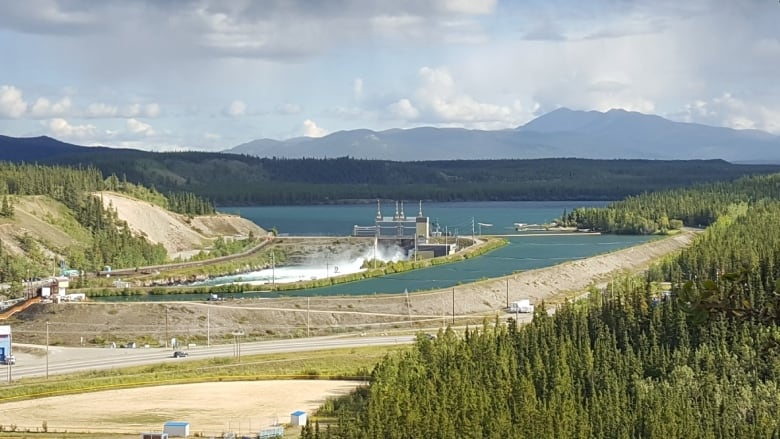 The headstock of the Whitehorse dam in front of Schwatka Lake. Water is rushing through the spillway. The scene is surrounded by boreal forest.