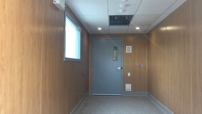 A grey metal door is shown at the end of a hallway lined with faux wood pannelling.