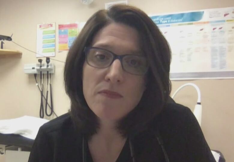 A woman with glasses and dark hair sits in a doctor's examination room.