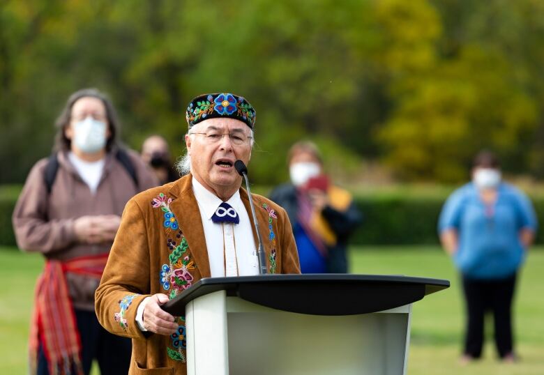 A man in a beaded hat speaks at a podium.