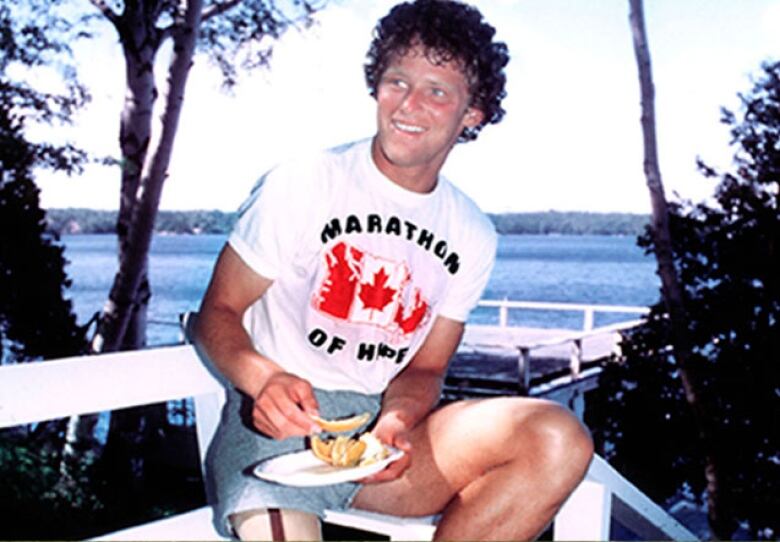 Terry  Fox eats some orange slices on a deck overlooking a body of water.  