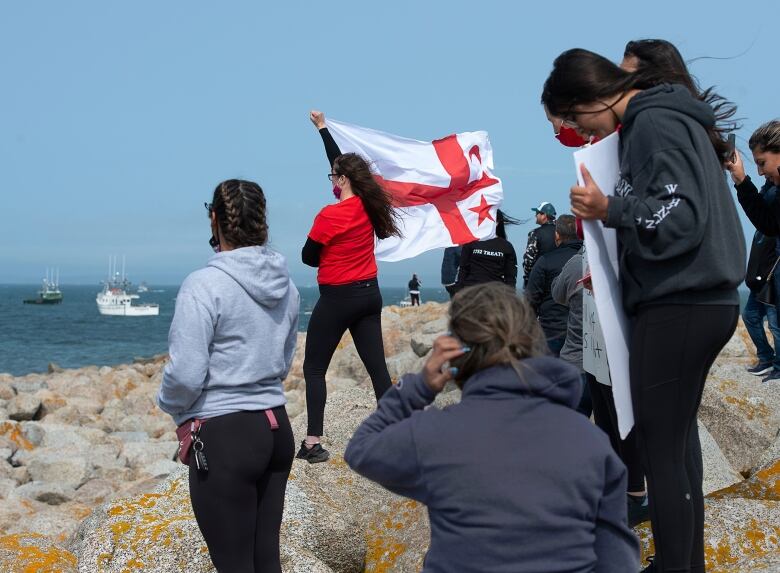 People stand on rocks overlooking the ocean, one of them holding a red and white flag.