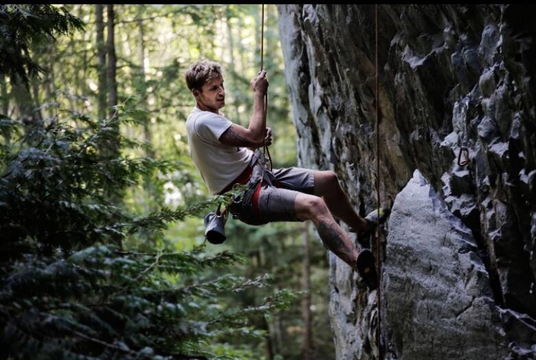 A man sits in climbing gear as he climbs a sheer rock wall in the forest.